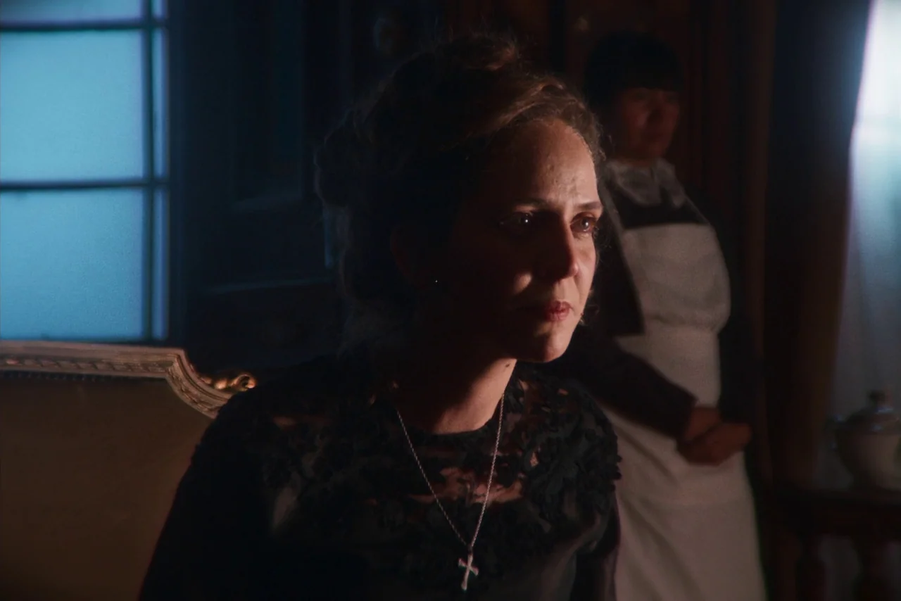 The Settlers (2023) movie still: medium — A dimly lit room with wood paneled walls features a woman sitting in the foreground. A maid in an apron…
