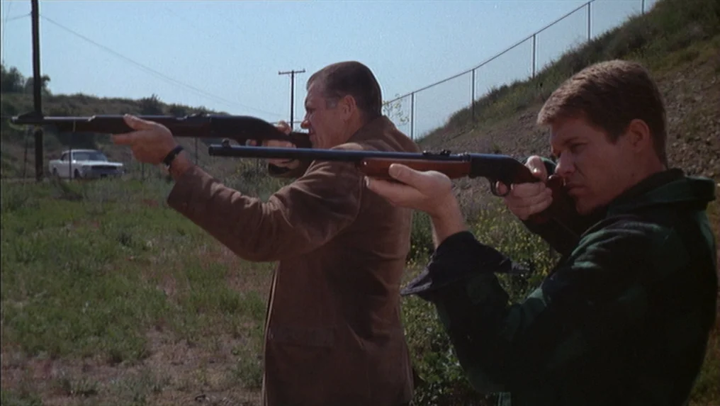 Targets (1968) movie still: medium — Two men stand in a grassy field against a chain-link fence on a slope. They hold hunting rifles and aim them…