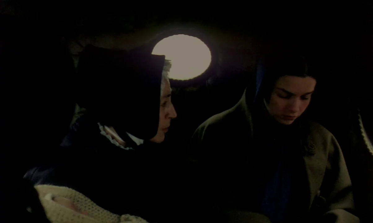Kamouraska (1973) movie still: medium — Two women in period clothing sit inside a dimly lit train carriage or coach. A circular window behind them…