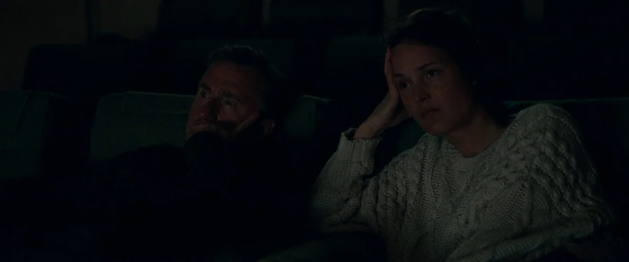 Bergman Island (2021) movie still: medium — A dark, dimly lit theater auditorium with rows of padded seats. A man and woman sit side by side…