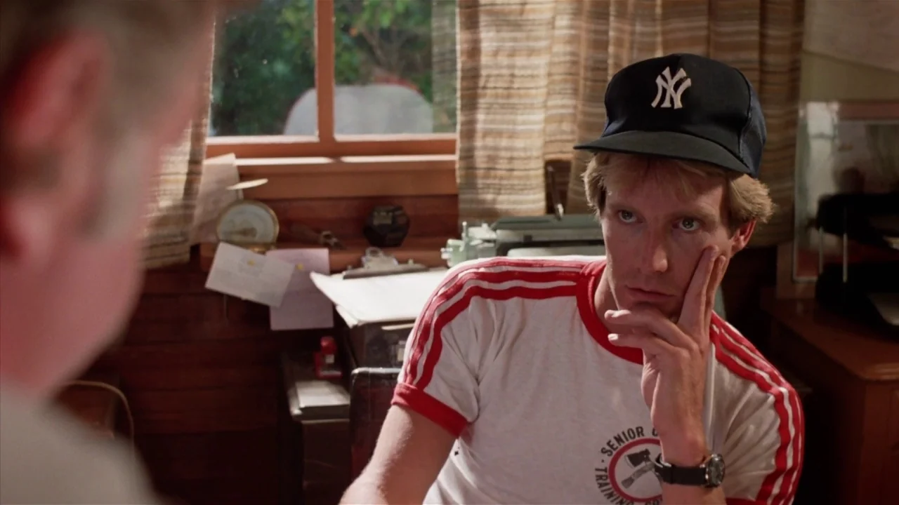 Friday the 13th Part 2 (1981) movie still: medium — A man sits at a cluttered desk in a space with wood-paneled walls. He wears a baseball cap and…
