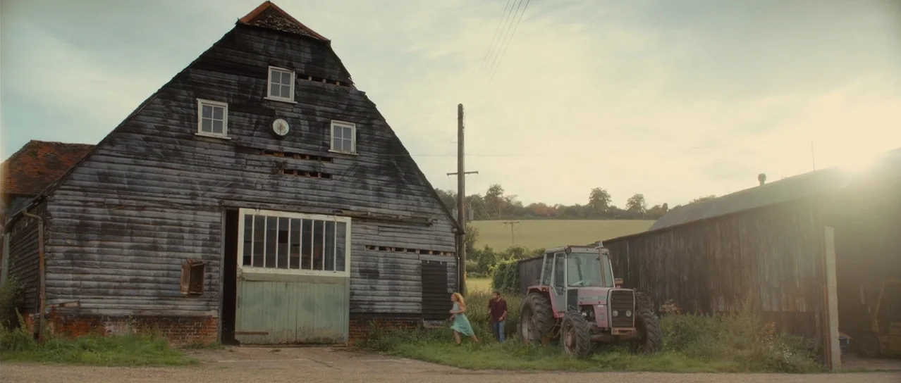 Road Games (2015) movie still: wide — A large weathered wooden barn stands beside a vintage red tractor in a field. Two people walk across the…