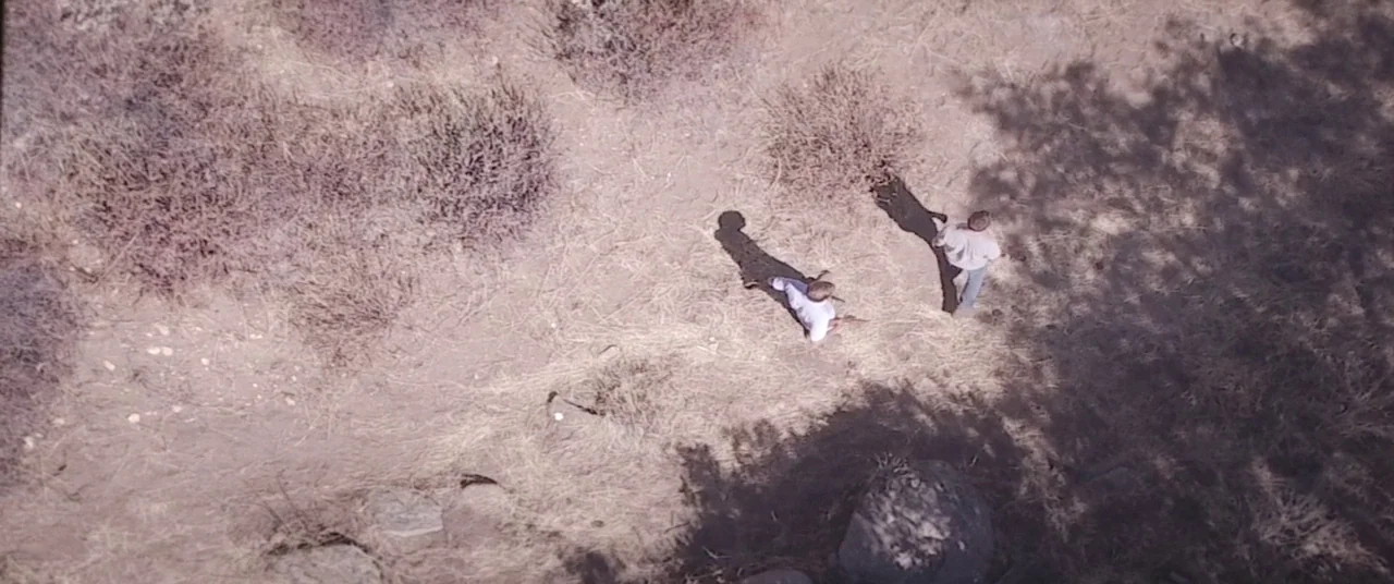 The Endless (2017) movie still: wide — A dry, sun-scorched field with sparse brush and patches of dead grass. Two men walk across the open, dusty…