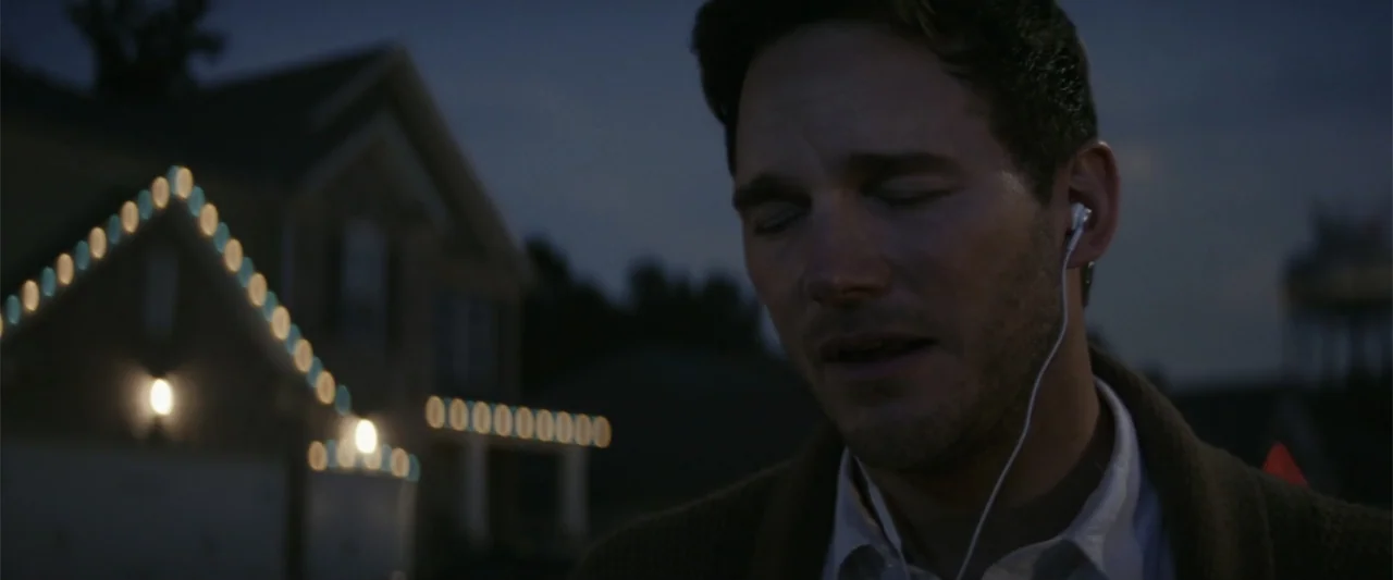 The Tomorrow War (2021) movie still: closeup — A man stands outdoors in the evening in front of a house at twilight. He wears white wired earphones…