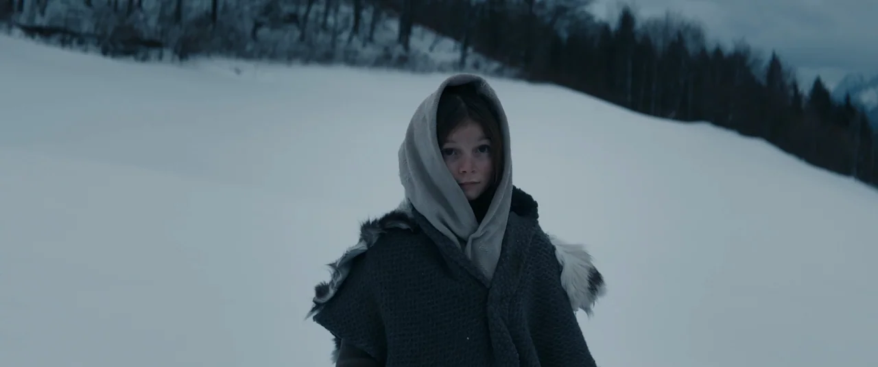 Hagazussa (2017) movie still: medium — A desolate snow-covered landscape stretches under a heavy, overcast winter sky. A young girl in a hooded grey…