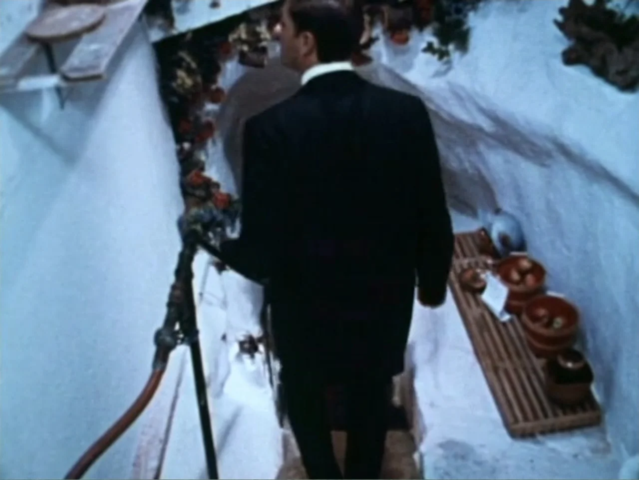 It's Alive (1974) movie still: medium — A narrow stone staircase with whitewashed walls features rustic decor and terracotta pottery bowls. A man in…