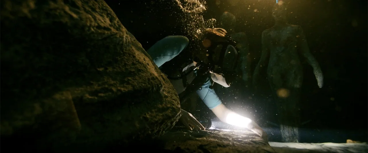 Anaconda (2024) movie still: medium — A dark underwater environment features a large stone rock and submerged human-like statues. A scuba diver…