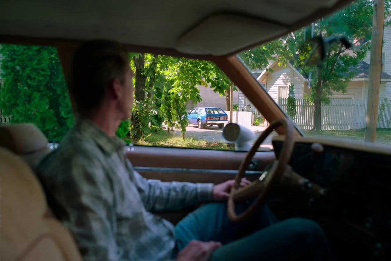 Barbarian (2022) movie still: medium — A view from inside a car looking toward a suburban street with houses. A man sits in the driver seat looking…