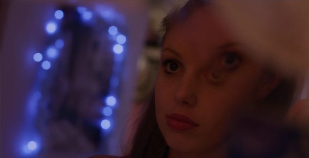 Dollhouse (2012) movie still: closeup — A dimly lit room at night features out-of-focus blue lights in the background. A young woman looks toward…