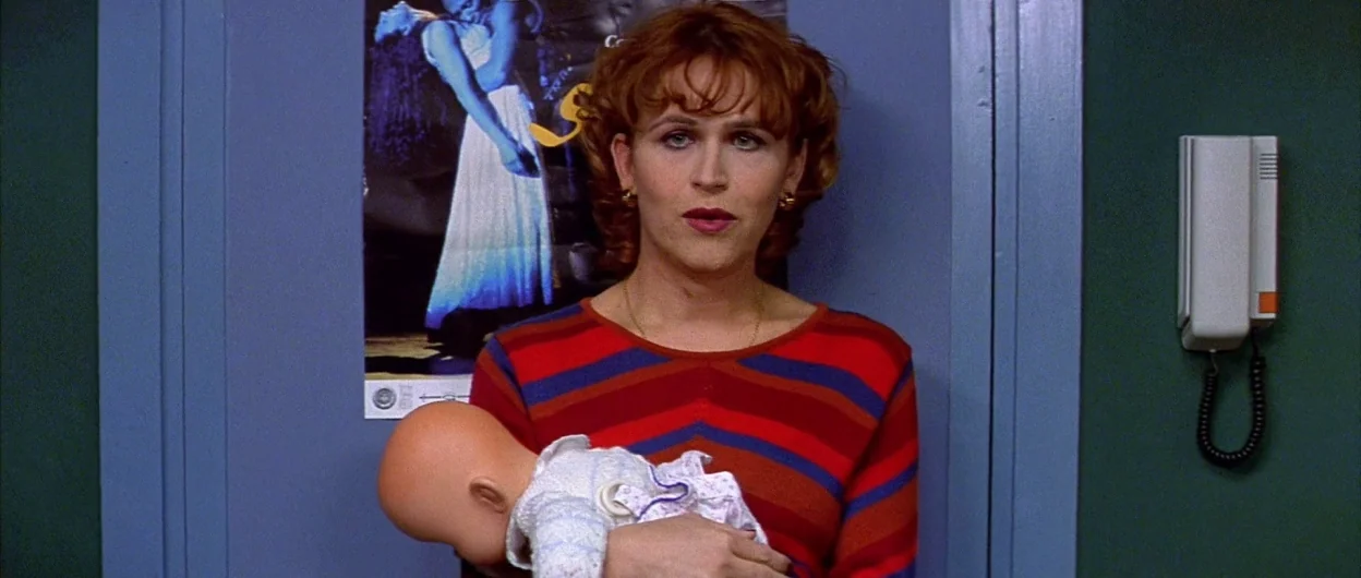 All About My Mother (1999) movie still: medium — A person stands before a light blue door with a poster and wall phone. The subject looks forward…
