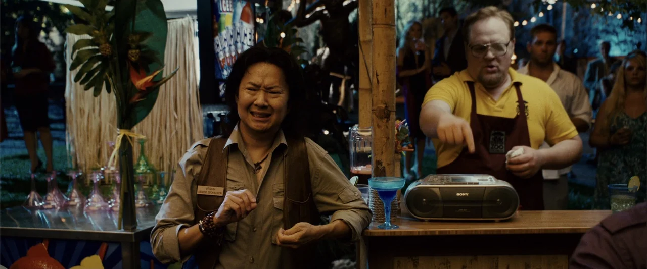 Zookeeper (2011) movie still: medium — An outdoor evening party features a bar setup with a wooden counter. A zookeeper stands beside a coworker who…