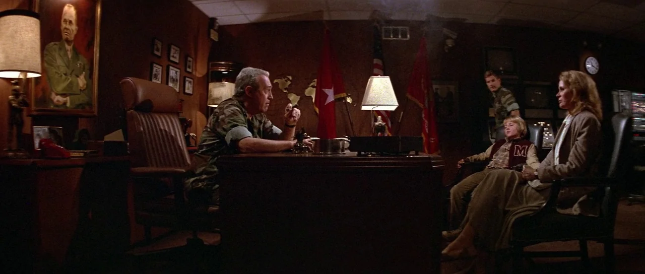 Invaders From Mars (1986) movie still: wide — A dimly lit room features a large wooden desk with flags behind it. A man speaks to an adult woman and…