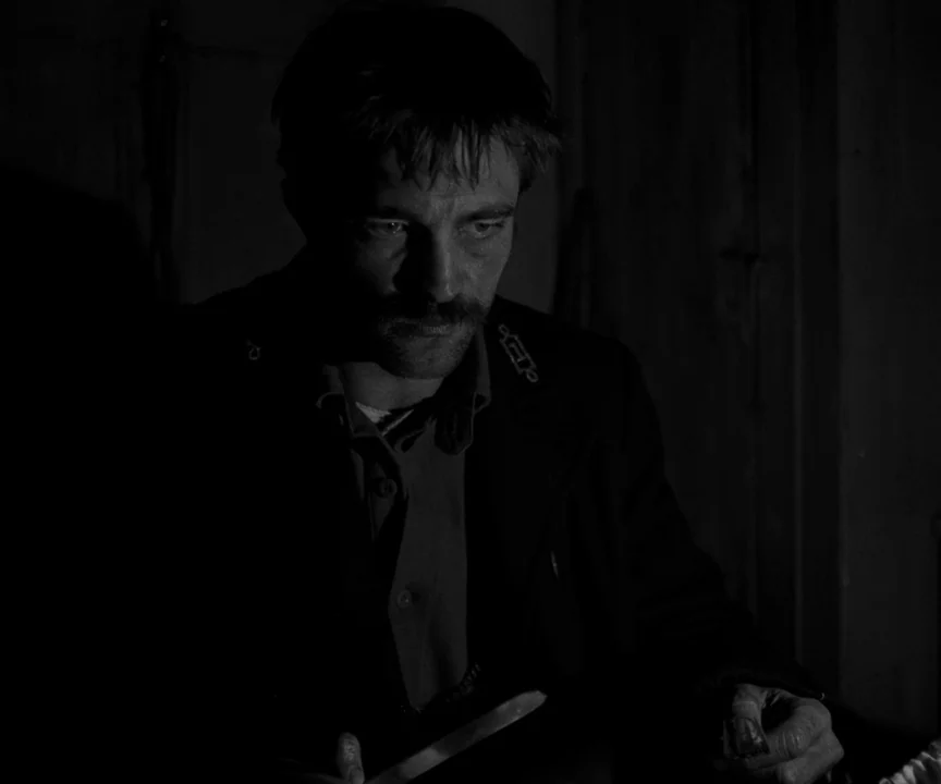 The Lighthouse (2019) movie still: medium — A dark, dimly lit room contains only a man sitting in the shadows. He inspects a small object while…