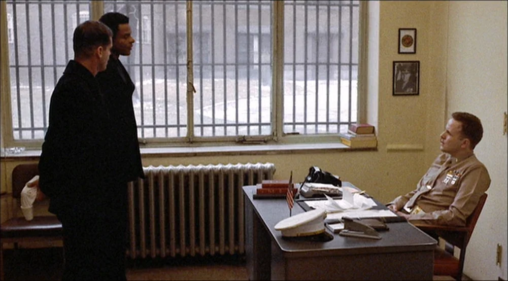 The Last Detail (1973) movie still: medium — An office features a desk, metal radiator, and barred windows showing an outdoor area. A seated officer…