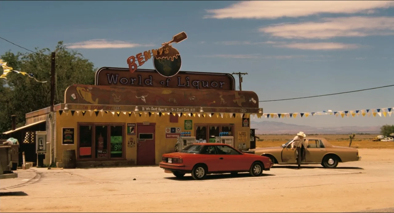 From Dusk Till Dawn (1996) movie still: wide — A yellow building with a prominent liquor sign sits in a vast desert landscape. A man in a cowboy hat…