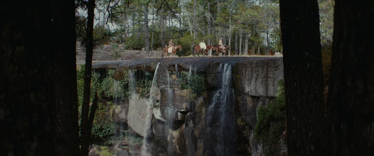 The Dead Don’t Hurt (2023) movie still: wide — A tall rock cliff face features water trickling over its mossy, textured edge. Three riders on brown…