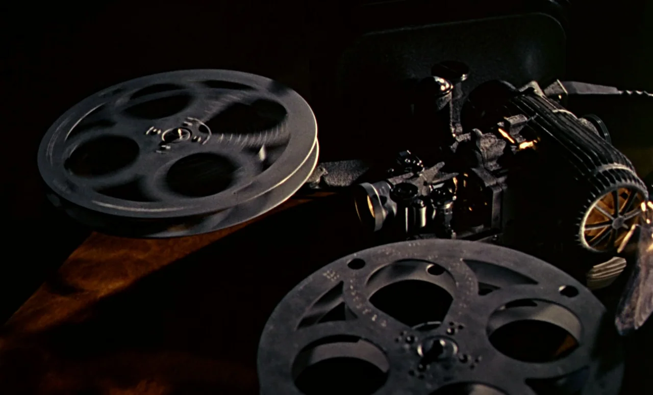 Peeping Tom (1960) movie still: closeup — A vintage mechanical film projector sits on a table with two film reels attached. One reel is in motion…