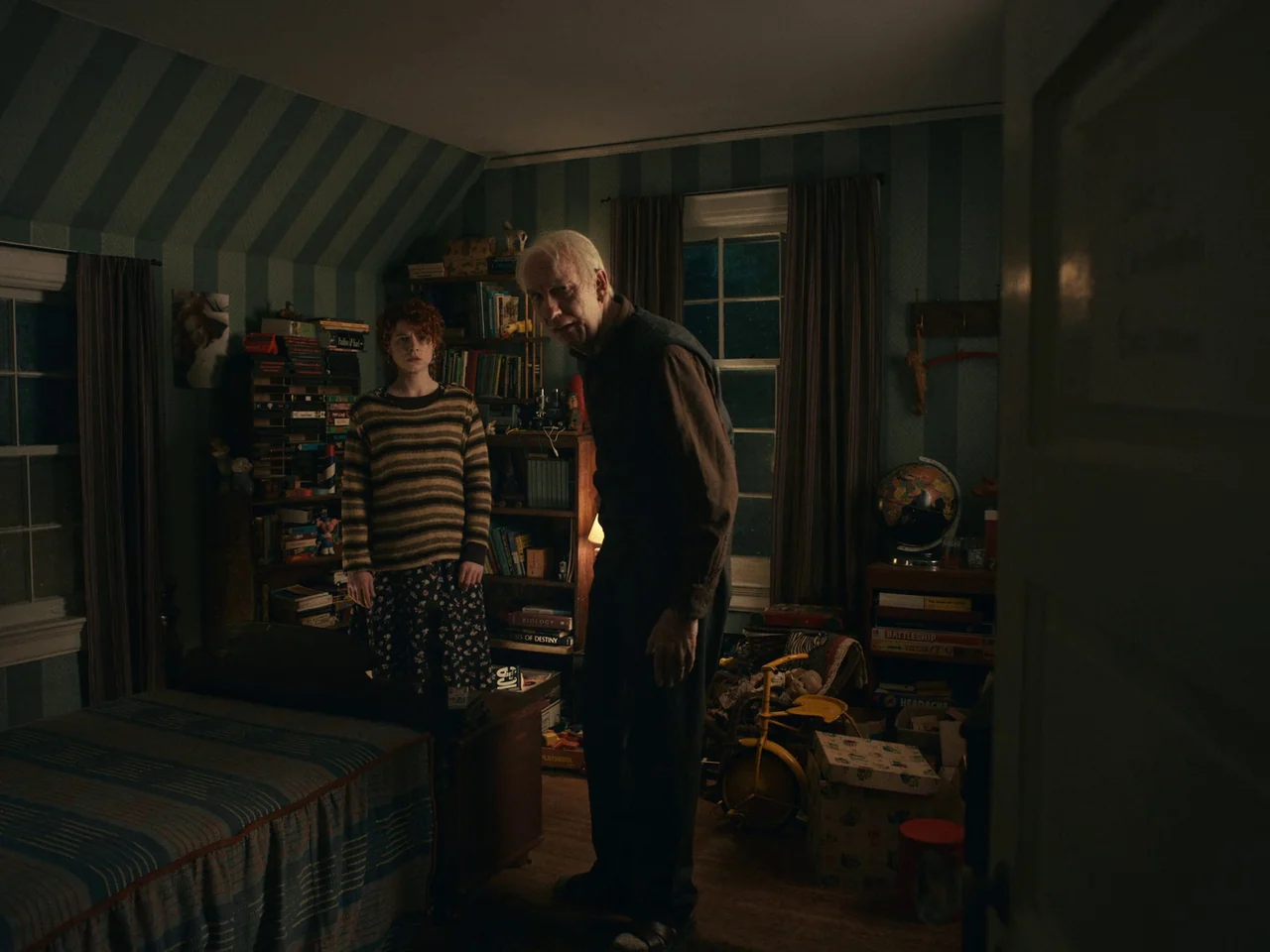 I’m Thinking of Ending Things (2020) movie still: fullbody — A cluttered room featuring blue striped wallpaper and shelves overflowing with books…