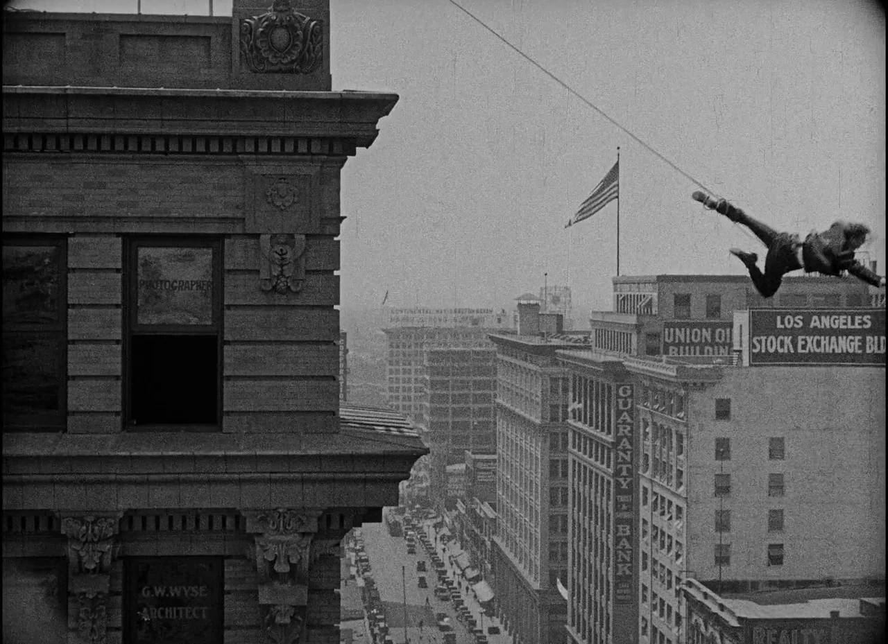 Safety Last! (1923) movie still: wide — A dense urban environment featuring early 20th-century commercial buildings and a visible American flag. A…