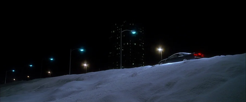 Kill Bill Vol. 1 (2004) movie still: wide — A dark car sits atop a snow-covered embankment under a row of streetlamps. A tall building with…