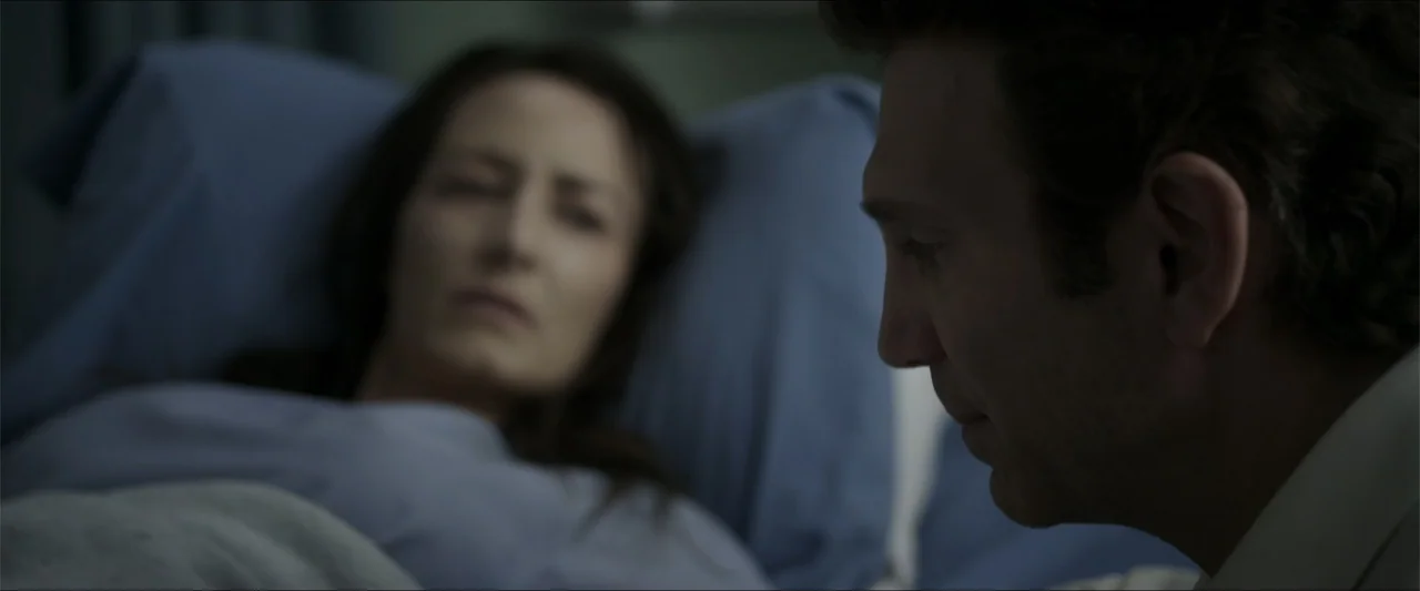 Heavenly Deposit (2019) movie still: medium — A dimly lit hospital room features a woman resting in a bed with blue linens. A man stands in the…