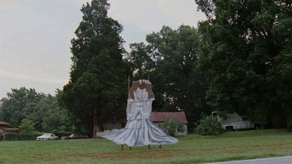 Junebug (2005) movie still: wide — A rural roadside landscape features a large, hand-painted wooden cutout of a girl. Dense green trees and a small…