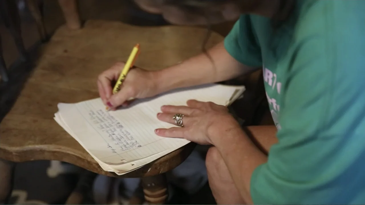 Meth Storm (2017) movie still: closeup — A person sits on a wooden chair writing on a piece of lined notebook paper. The subject is wearing a green…
