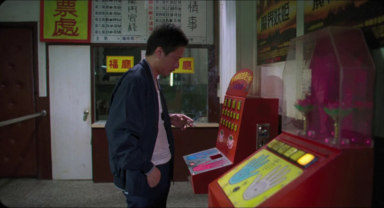Goodbye, Dragon Inn (2003) movie still: medium — A dimly lit lobby features posters on walls and a vintage red gaming kiosk. A man stands alone in…