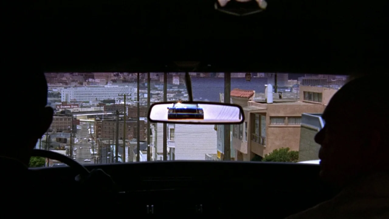 Bullitt (1968) movie still: medium — The interior of a car looking out onto a steep city street. A dark vehicle is reflected in the rearview mirror.