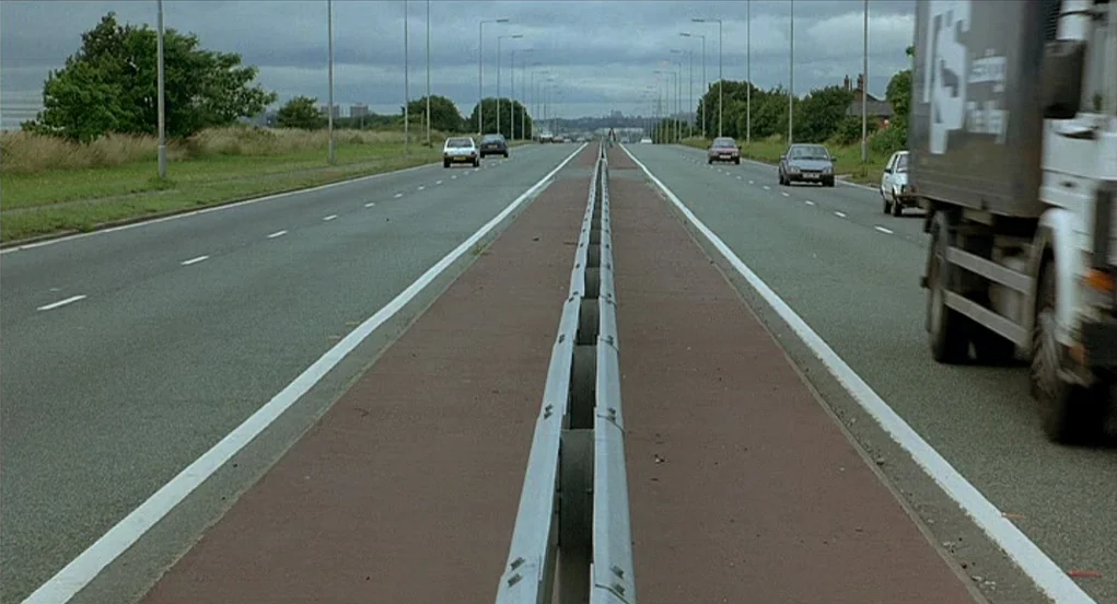 Butterfly Kiss (1995) movie still: establishing — A wide, divided asphalt highway stretches into the distance under an overcast sky. Several…