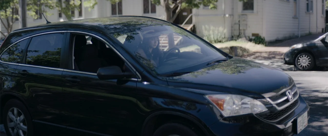 A Christmas Mystery (2022) movie still: medium — A residential street features a dark Honda SUV parked near a white building. A woman sits in the…