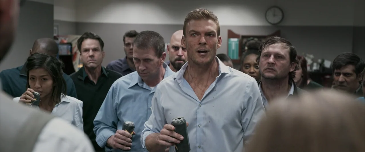 Office Uprising (2018) movie still: medium — A crowded office breakroom with a clock on the wall and various employees standing. A man at the center…
