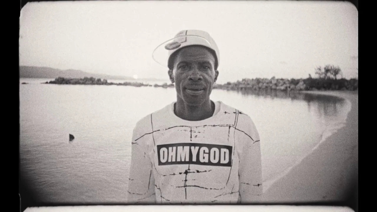 Black Mother (2018) movie still: medium — A man stands before a calm body of water with a shoreline stretching behind. He wears a white shirt and…