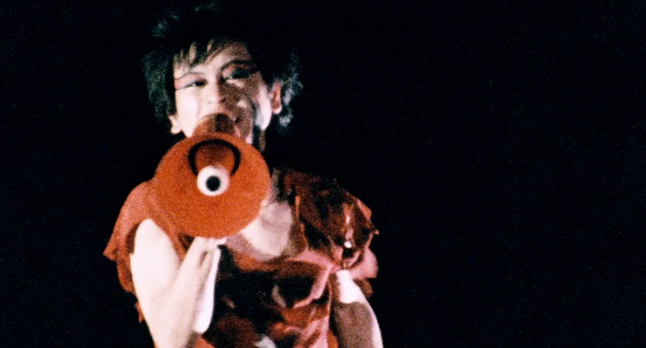 Burst City (1982) movie still: medium — A performer stands on a dark stage against a completely black background. The person shouts into a red…