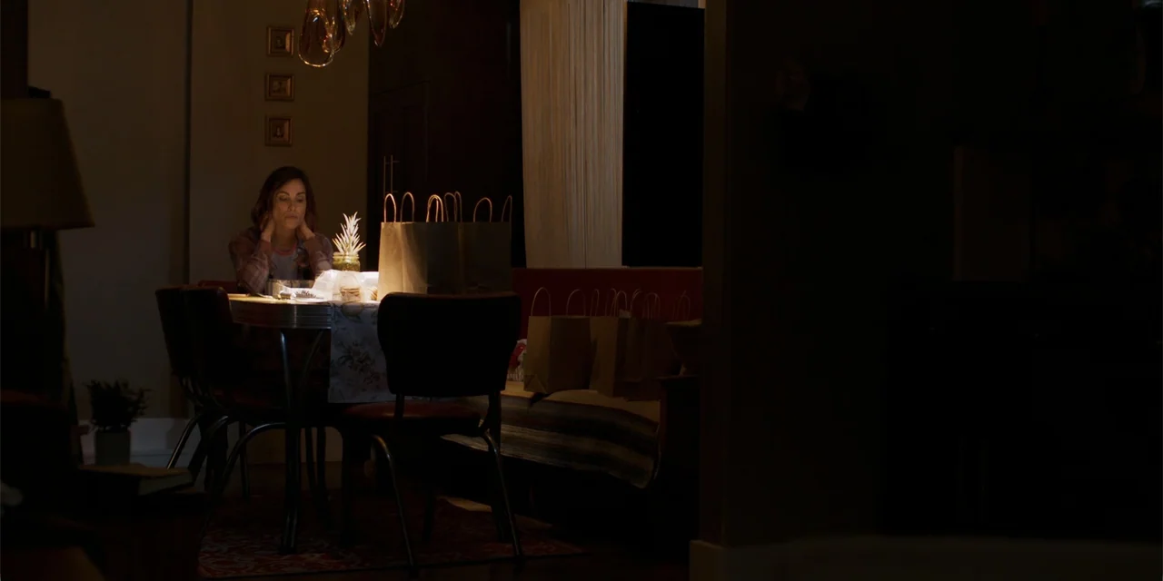 Demonic (2015) movie still: medium — A dimly lit dining room contains a wooden table and several paper shopping bags. A woman sits alone at the…