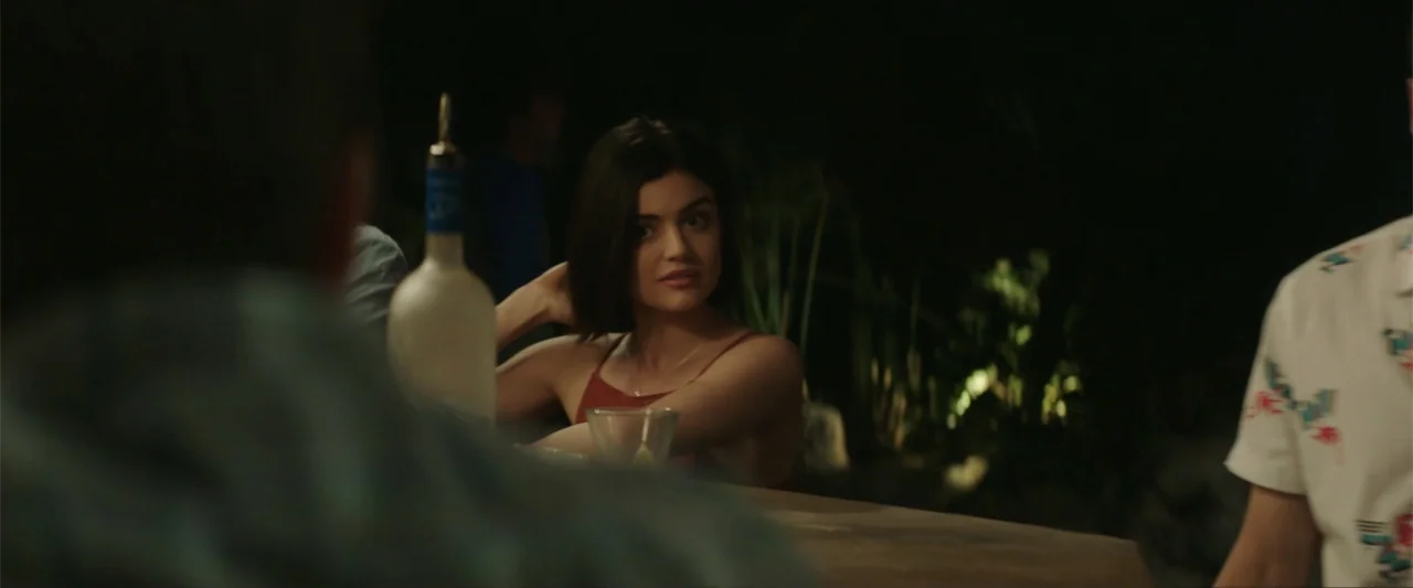 Truth or Dare (2018) movie still: medium — A woman sits at an outdoor bar counter at night with blurred foliage behind. She rests her hand on the…