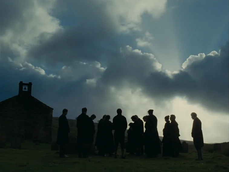 Wuthering Heights movie still: wide — A group of people stands in a circle in a rural moorland cemetery. An old stone building with a small bell…