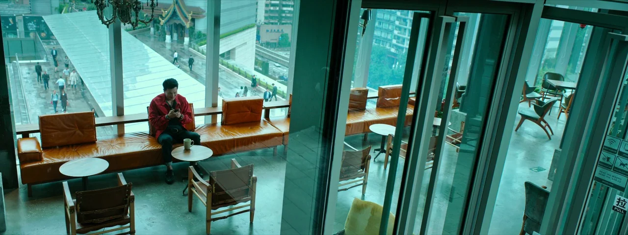 Under The Light (2023) movie still: wide — A modern cafe with floor-to-ceiling windows overlooks a wet urban plaza. A man in a red jacket sits alone…