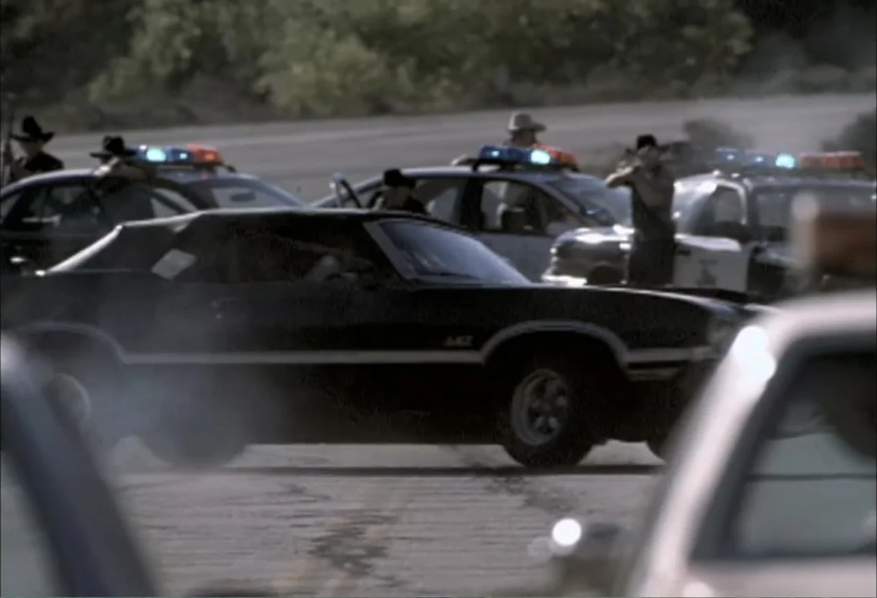 Black Cat Run (1998) movie still: wide — A black vintage muscle car drives along a paved road surrounded by police cruisers. Several officers…