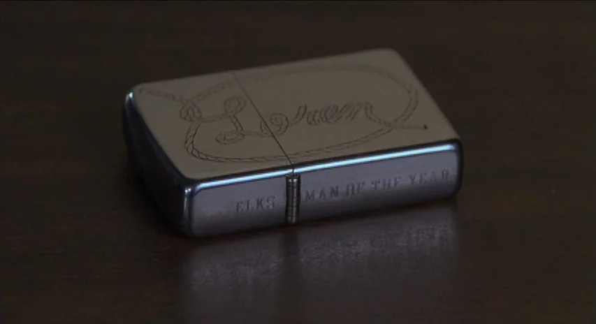 Blood Simple (1984) movie still: closeup — A silver metal lighter sits on a polished dark wood table surface. The lighter features an engraving of a…