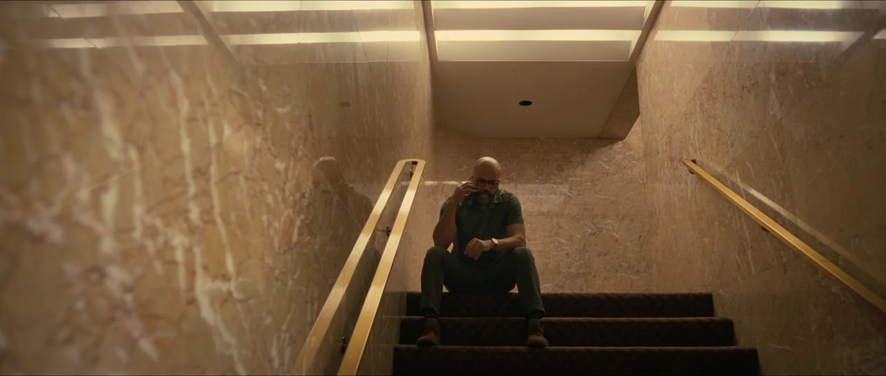 American Fiction (2023) movie still: medium — A man sits on stairs in a hallway lined with polished brown marble panels. The subject rests on the…
