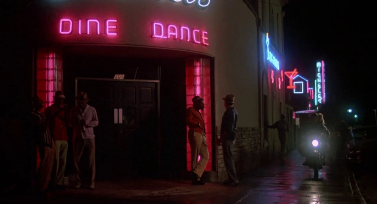 Peggy Sue Got Married (1986) movie still: wide — An exterior street scene at night features a neon-lit club entrance with glass blocks. Several men…