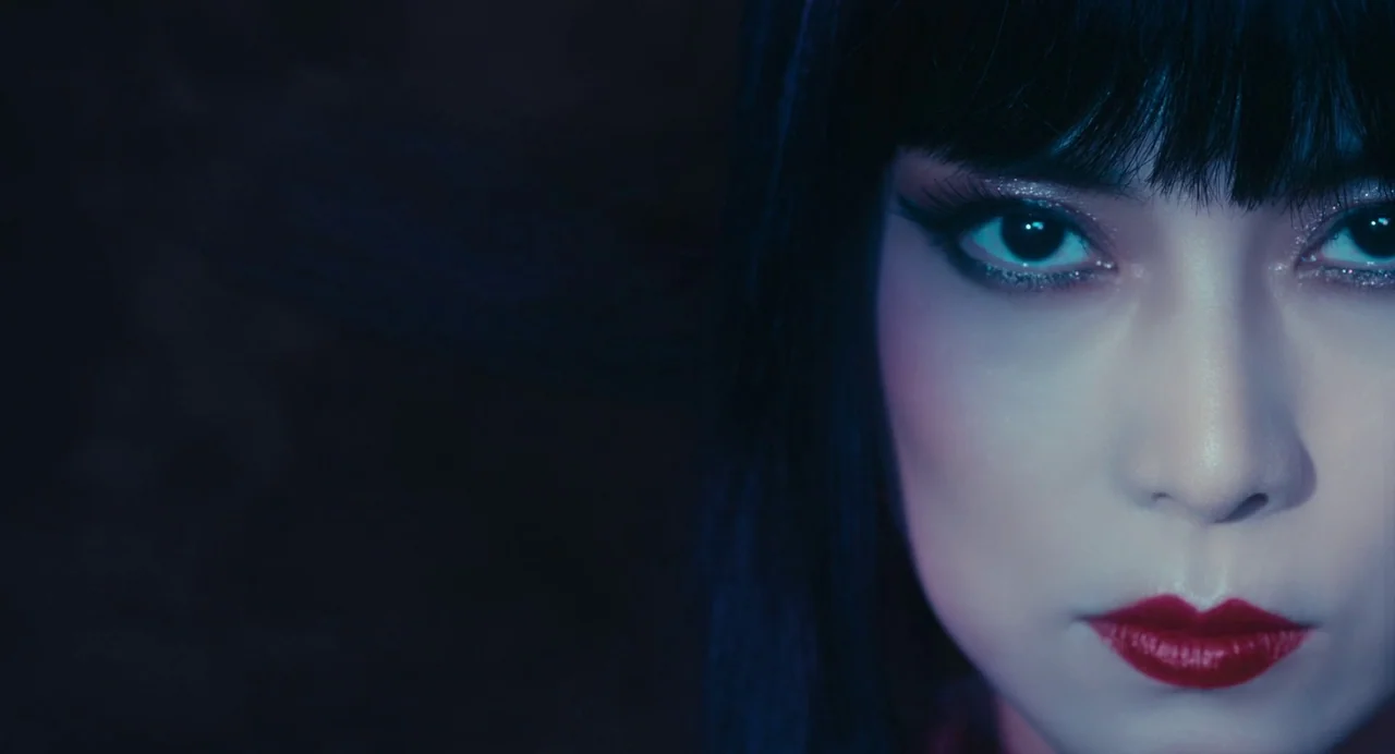 xxxHolic (2022) movie still: closeup — A woman's face is illuminated from the right side against a dark, featureless backdrop. Her eyes are centered…