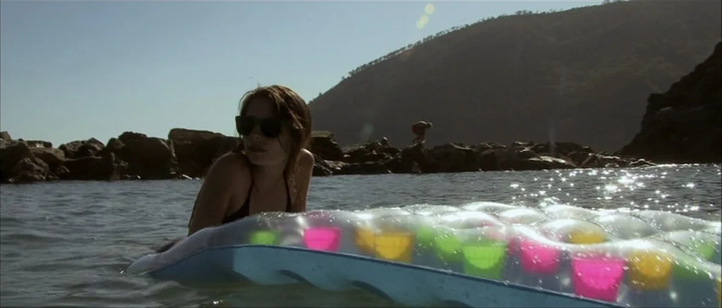 Genova (2008) movie still: medium — A rocky coastal cove features a person floating on a multicolored inflatable air mattress. The individual looks…