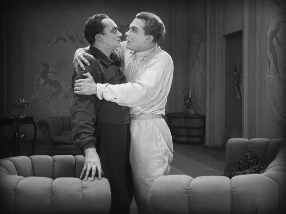 Metropolis (1927) movie still: medium — A room with art deco murals on the walls and upholstered furniture. Two men face each other while one grips…