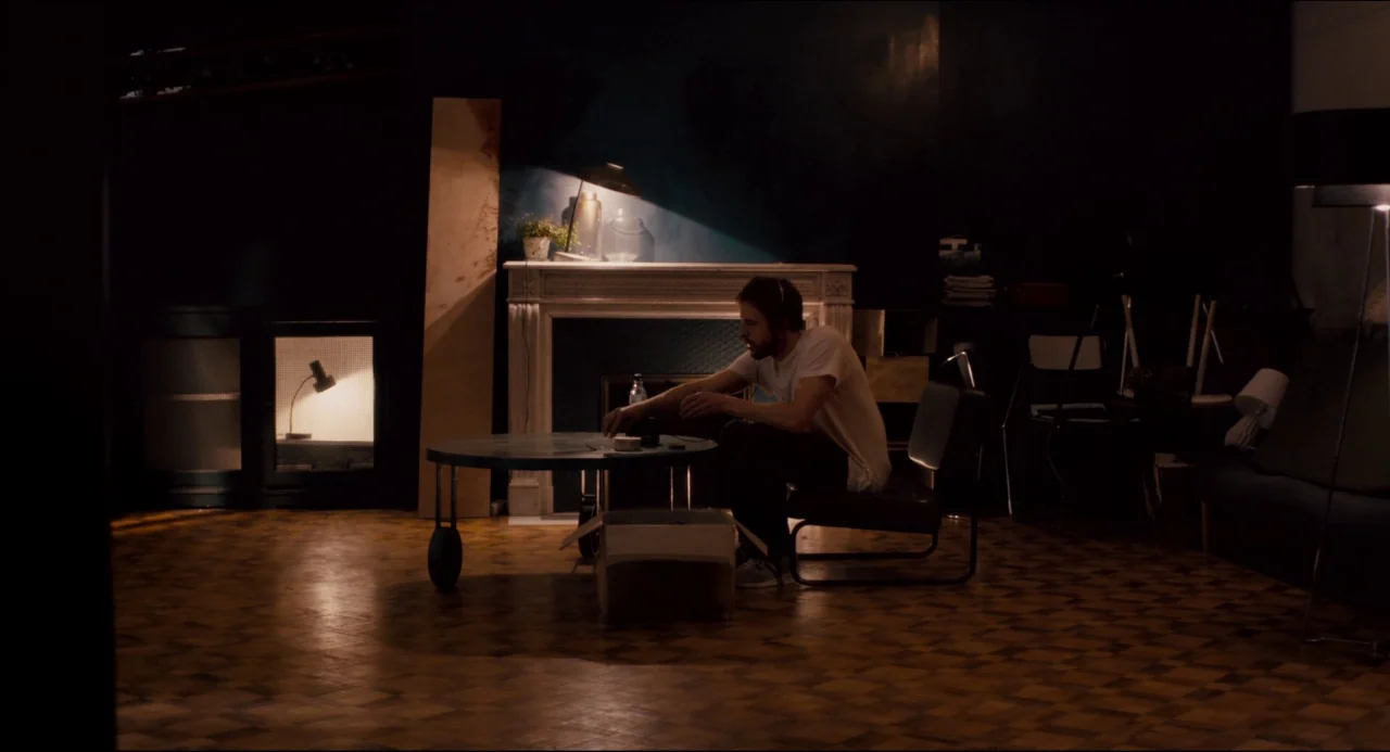 The Night Eats the World (2018) movie still: wide — A dimly lit living room features a parquet floor and a classic fireplace mantel. A lone man sits…