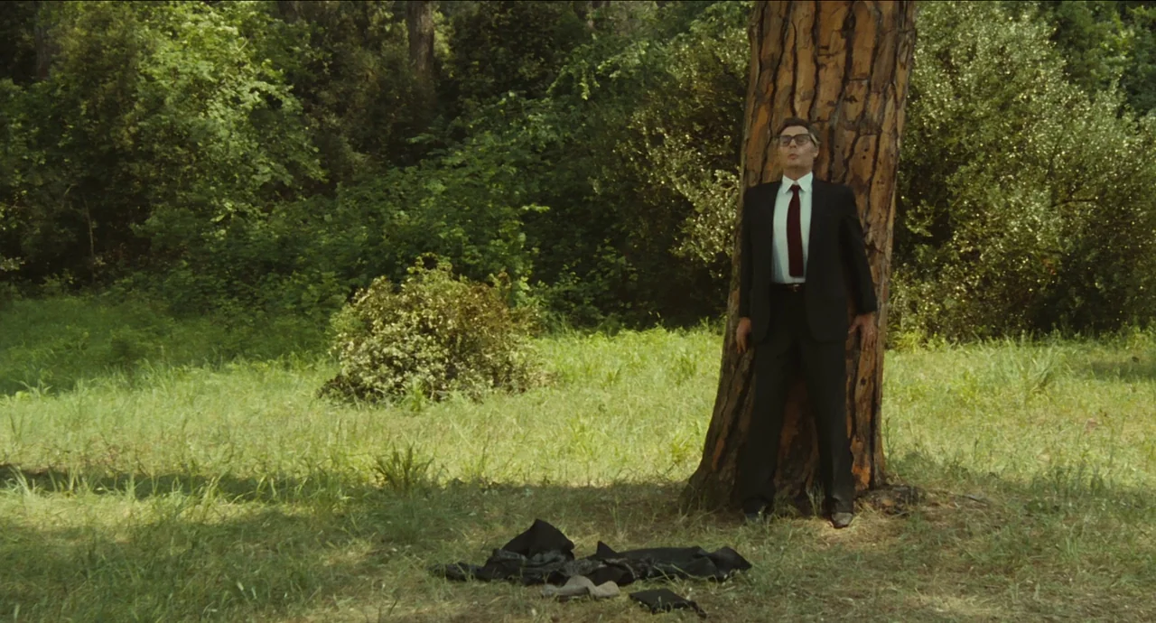 City of Women (1980) movie still: fullbody — A man in a suit stands against a large pine tree in a meadow. A discarded jacket lies on the grass at…