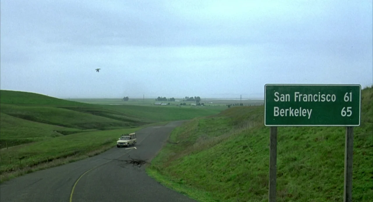 Hulk (2003) movie still: wide — A rural asphalt road winds through rolling green hills under a flat overcast sky. A single car drives along the road…