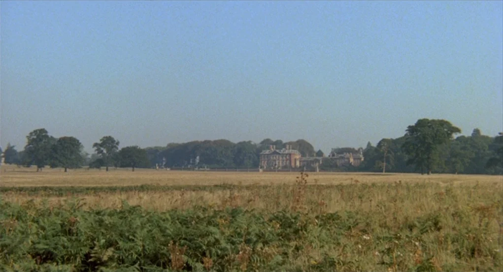 The Go-Between (1971) movie still: establishing — A wide expanse of golden-brown field stretches toward a distant historic stone mansion. Mature…