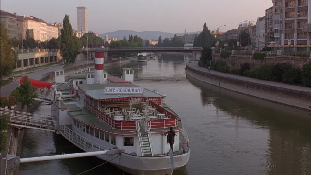 Before Sunrise (1995) movie still: establishing — A river canal flows through a European city lined with mid-century residential buildings. A docked…